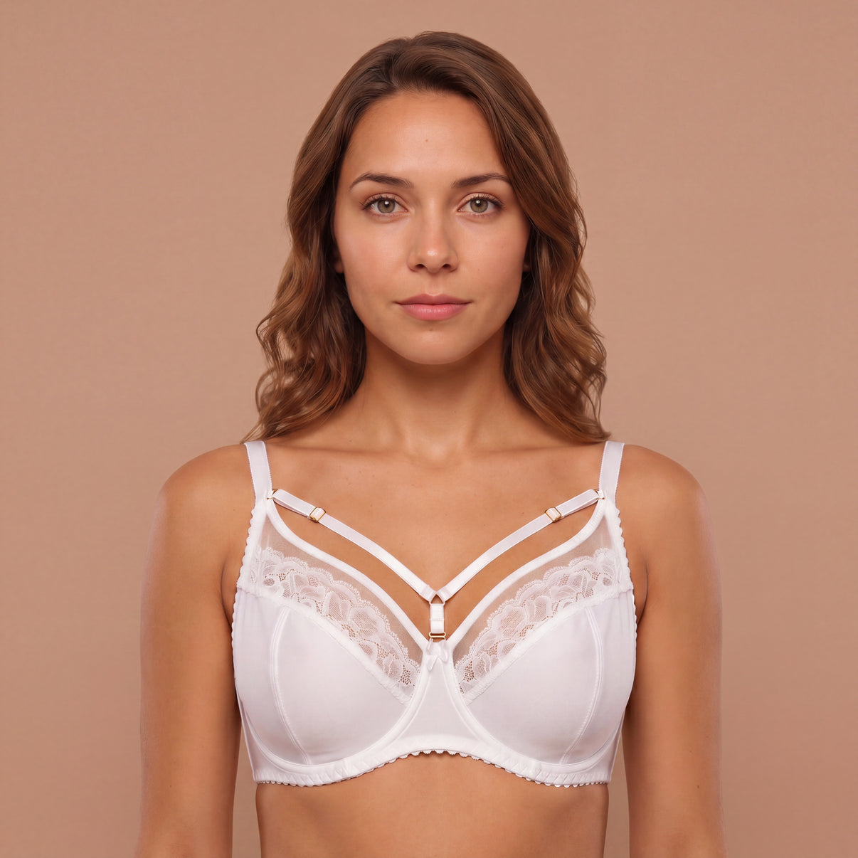 A woman modeling a white lace-trimmed bralette with thin adjustable straps and a small gold-toned center accent, photographed against a plain warm beige studio background.